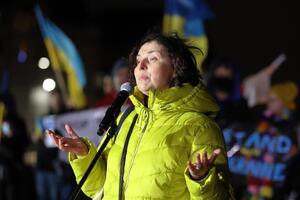 Kobieta w średnim wieku, ubrana w żółtą kurtkę przemawia do uczestników manifestacji. Widzimy jej lewy profil, mówiąc - rozkłada ręce. W tle widać uczestników manifestacji i trzymane przez nich ukraińskie flagi