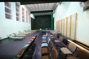 na zdjęciu sala gimnastyczna w jasnych zielonych kolorach ściany, widać kilkadziesiąt ustawionych krzeseł, w tle rozłożono kilka łóżek polowych