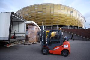 Sztaplarka przewozi kartony zapakowane, obok tył otwarty ciężarówki, w tle fasada stadionu Polsat Plus Arena Gdańsk