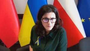 Kobieta w okularach, siedzi przy stole, ma przypiętą do bluzki kokardę w barwach Ukrainy. Z tyłu 