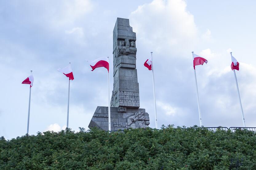 wysoki, kamienny pomnik w stoi na szczycie wzgórza otoczony masztami, na których powiewają biało-czerwone flagi