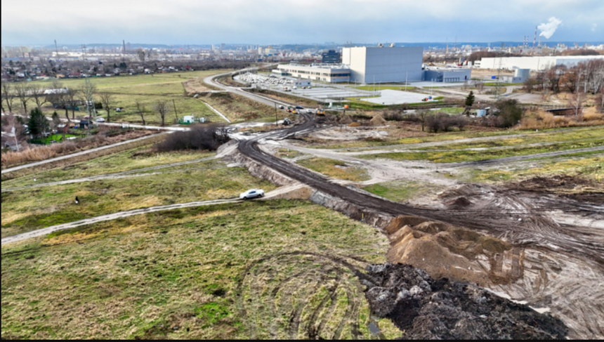 zdjęcie z drona, widać zielony teren, częściowo rozkopany przez sprzęt budowlany, w tle budynek jednej z fabryk
