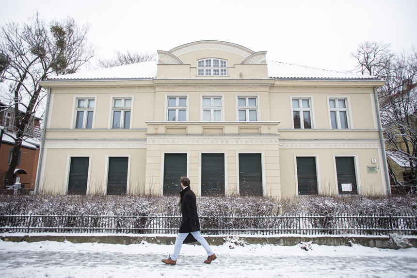 mężczyzna przechodzi obok zabytkowego dwukondygnacyjnego budynku, cała okolica pokryta jest śniegiem.