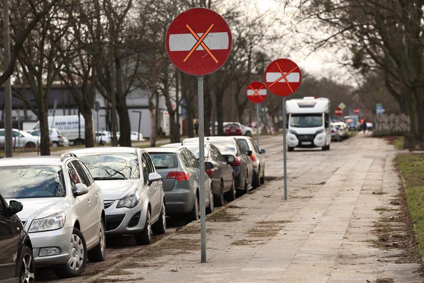 na zdjęciu chodnik przy którym wzdłuż zaparkowanych jest kilka samochodów osobowych, widać też kilka znaków drogowych informujących o zakazie wjazdu
