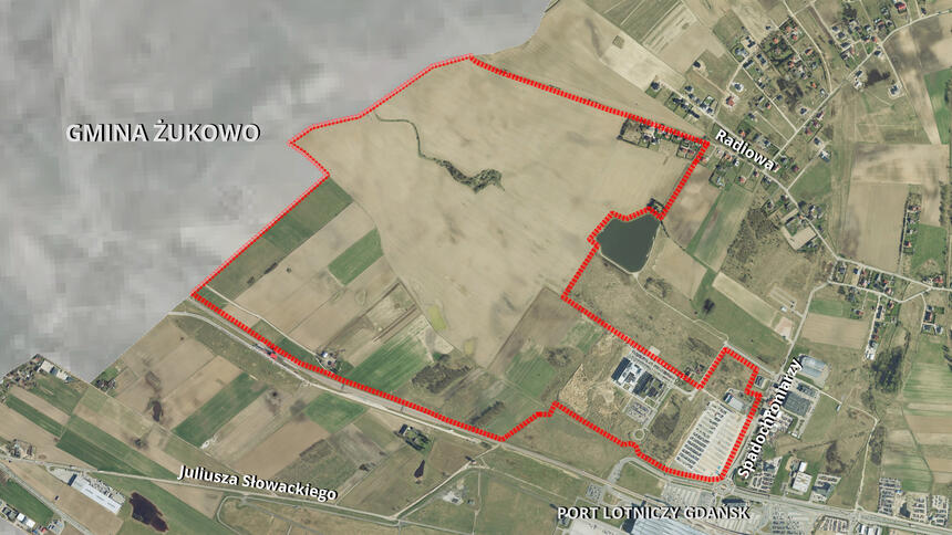 Zdjęcie przedstawia widok z lotu ptaka na określony obszar zaznaczony czerwoną linia przerywaną. Na dole i w górnej części obrazu znajdują się nazwy miejsc, takie jak "Juliusza Słowackiego" i "Radiowa". Wzdłuż dolnej krawędzi zdjęcia widoczna jest nazwa "Port Lotniczy Gdańsk", na lewej stronie widoczny jest napis "GMINA ŻUKOWO", co sugeruje, że ten obszar należy do określonej jednostki administracyjnej w Polsce. 