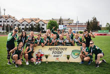 Grupa zawodniczek rugby pozuje do zdjęcia po zwycięskim meczu, który zapewnił im mistrzostwo Polski. Kobiety są uśmiechnięte i pokazują palcem numer 1. Między nimi stoi baner z napisem "Mistrzynie Polski rugby 7" oraz złoty puchar.