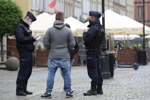 Na zdjęciu widać dwóch policjantów w służbowych granatowych mundurach. Legitymują oni mężczyznę ubranego w szarą bluzę i niebieskie dżinsy. W tle widać kremowe parasole umieszczone w jednym z ogródków gastronomicznych