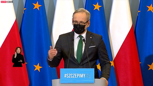 Mężczyzna w srednim wieku, w masecze, prze mikrofonem, w tle flagi Polski i UE 