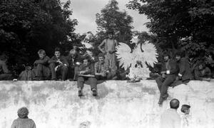 mężczyźni siedzą na wysokim murze, obok nich ustawiony duży herb państwa - orzeł w koronie, kilka osób z dołu patrzy na nich