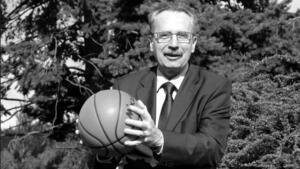 Czarno-białe zdjęcie: uśmiechnięty mężczyzna w średnim wieku w garniturze i okularach, w dłoniach trzyma piłkę do koszykówki