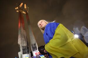 pomnik poległych stoczniowców (trzy krzyże) podświetlony na kolory wolnej Ukrainy: biało i czerwono, pod pomnikiem stoi dziewczyna z blond włosami otulona flagą Ukrainy