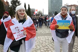 Ludzie podczas marszu. Na pierwszym planie (po lewej) młoda kobieta z nałożoną na ramiona flagą Białorusi, w dłoni trzyma kartkę z czerwonym napisem: FREE BIELARUS oraz młody mężczyzna (po prawej) z narzuconą na ramiona flagą Białorusi a w dłoniach trzyma niebiesko-żółtą kartkę z napisem: STOP PUIN, STOP WAR 