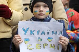 na zdjęciu chłopiec ze smutną miną, ma czarną czapkę na głowie, na obu policzkach wymalowaną żółto-niebieską flagę ukrainy, w rękach trzyma kartkę z napisem w języku ukraińskim