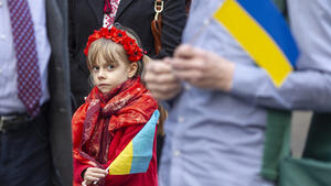 Mała dziewczynka ubrana w czerwony strój, z przepaską na czole, w ręku trzyma ukraińską flagę