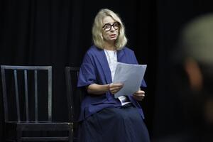 sala teatralna, na krześle siedzi kobieta, blondynka w okularach, czyta z trzymanych w dłoniach kartek