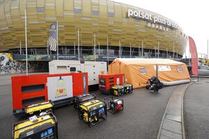 na zdjęciu kilka agregatów prądu, po prawej widać duży namiot pomarańczowy przy którym kuca właśnie fotoreporter, w tle bryła gdańskiego stadionu