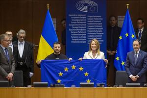 Pośrodku zdjęcia stoją mężczyzna i kobieta (z prawej) za stołem prezydialnym. Wspólnie pozują do zdjęcia z flagą Unii Europejskiej, którą razem trzymają w rękach. Po prawej i lewej, na marginesach zdjęcia widać kilka innych osób