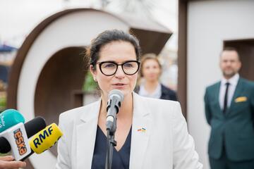 Kobieta w okularach w białej garsonce przed mikrofonami. W tle niewyraźnie dwie inne osoby 