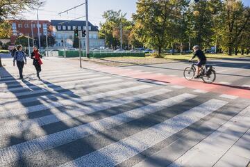 na zdjęciu widać pasy przejścia dla pieszych, obok biegnie droga rowerowa o czerwonej nawierzchni, jedzie nią rowerzysta, po pasach idą dwie osoby