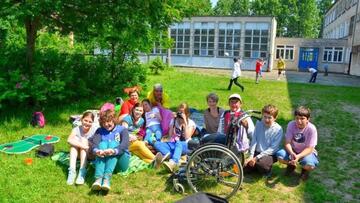 Grupa dzieci siedząca na kocu na trawniku przed szkołą, obok stoi wózek inwalidzki