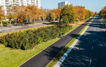 osiedle bloków wśród jesiennych drzew, słoneczna pogoda wzdłuż ulicy biegnie świeżą, czarna asfaltowa droga rowerowa, po której jedzie rowerzysta
