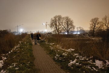 zdjęcie nocne, chodnikiem wzdłuż bastionów idzie kilka osób z psem, w tle oświetlone dźwigi budowalne i budynki oraz drzewa 