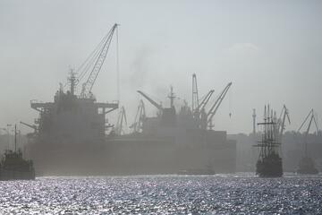 Żaglowiec płynie przez port, w tle widoczne statki zacumowane przy nabrzeżu w powietrzu widoczna ciemna chmura ciemnego pyłu.