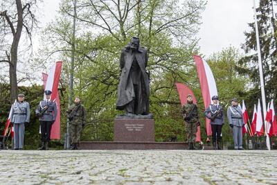 Zdjęcie przedstawia pomnik Józefa Piłsudskiego podczas jednej z gdańskich uroczystości. Pomnik stoi centralnie, na prawo i lewo od niego - po kilka osób. Obok, z jednej i drugiej strony - biało-czerwone flagi 
