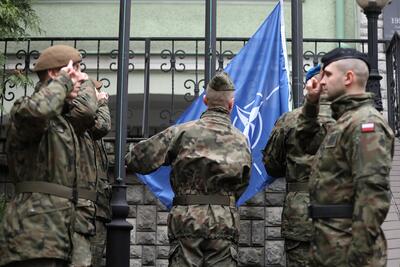 Żołnierz wciąga na maszt flagę NATO. Po jego prawej i lewej stronie stoją żołnierze, którzy salutują 