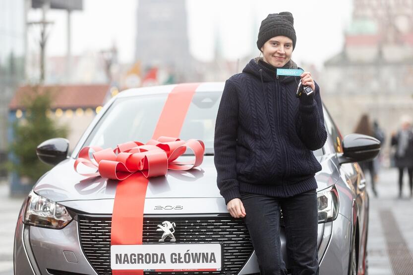 na zdjęciu młoda szczupła kobieta, opiera się delikatnie o stojący za nią samochód marki peugeot w kolorze szarym z czerwoną kokardą jaką stosuje się przy prezentach, kobieta trzyma w ręku kluczyki do samochodu