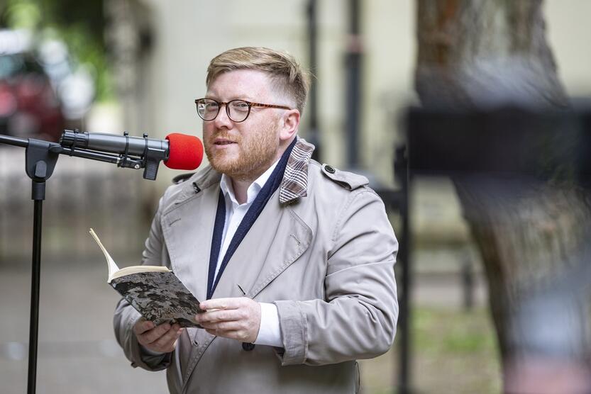 niewysoki mężczyzna stoi przy mikrofonie, trzyma w dłoniach tomik poezji, czyta