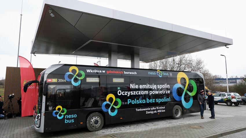 Na zdjęciu widać czarny autobus elektryczny z grafiką i napisami po polsku, stojący pod zadaszoną stacją ładowania. Na boku autobusu widoczne są hasła promujące czystą energię i transport bez emisji spalin, takie jak "Nie emituje spalin Oczyszczam powietrze" i "Polska bez spalin". Jest także informacja o krótkim czasie tankowania (ładowania) tylko 15 minut i zasięgu 450 km. Z przodu autobusu znajduje się logo firmy, które składa się z trzech splatających się pierścieni w różnych kolorach. Dwie osoby stoją obok autobusu i prowadzą rozmowę. W tle widoczna jest budowa lub plac przemysłowy, a także flaga, prawdopodobnie promująca wydarzenie lub produkt związany z autobusem. Całość odbywa się w ciągu dnia, a niebo jest pochmurne