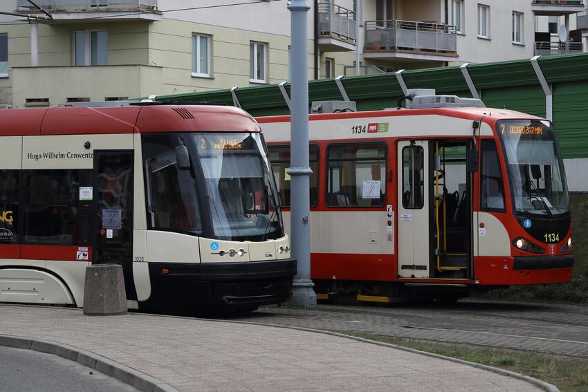 Na zdjęciu widać dwa tramwaje. Pierwszy z nich, w kolorze czerwono-kremowym, ma otwarte drzwi i znajduje się na pierwszym planie. Wygląda na starszy model. Drugi tramwaj, który wygląda na nowocześniejszy, ma ciemniejsze barwy z elementami czerwieni i beżu. Znajduje się za pierwszym tramwajem i wydaje się być zamknięty. Obie jednostki są zatrzymane na jakimś przystanku lub stacji tramwajowej. W tle widać mieszkalne bloki i trochę zieleni.