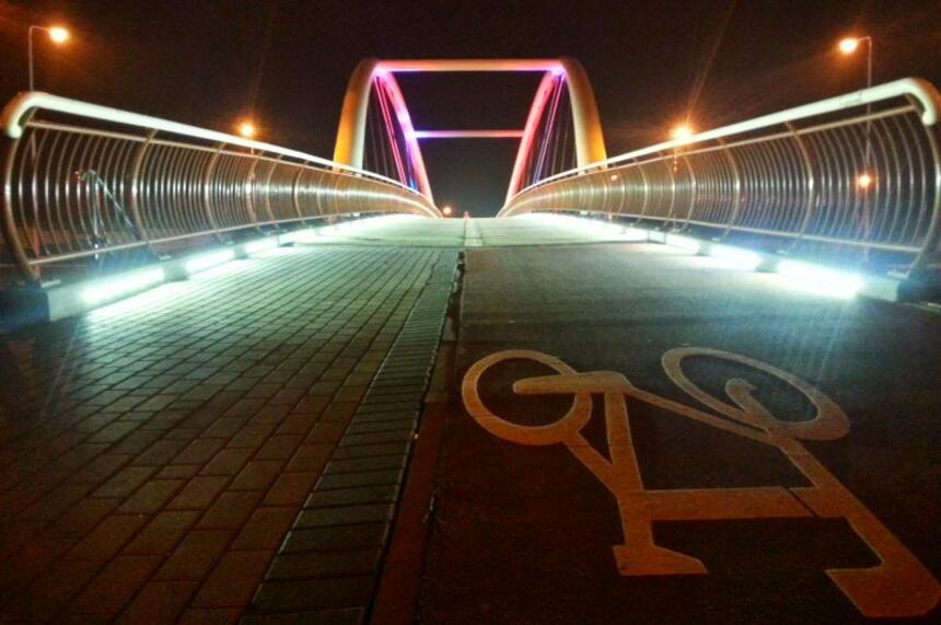 Na nocnym zdjęciu widać podświetloną kładkę pieszo-rowerową; chodnik dla pieszych ułożony jest z kostek, droga rowerowa z asfaltu, widać na niej biały symbol roweru.