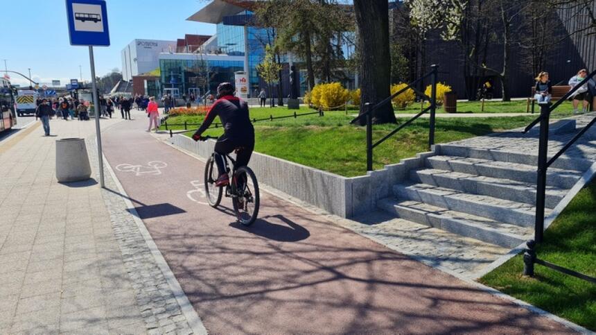 na zdjęciu fragment drogi rowerowej o czerwonej nawierzchni, jedzie po niej rowerzysta, po lewej widać fragment chodnika w kolorze szarym, po prawej widać nowe schody, kilkustopniowe, w tle budynki centrum handlowo-usługowego