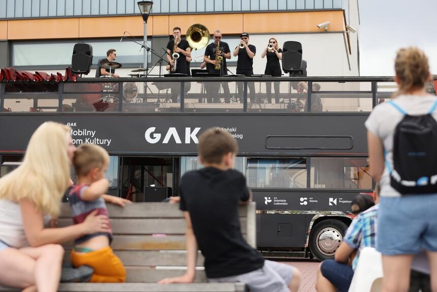 muzycy dęci na dachu autobusu grają koncert, na dole stoi publiczność