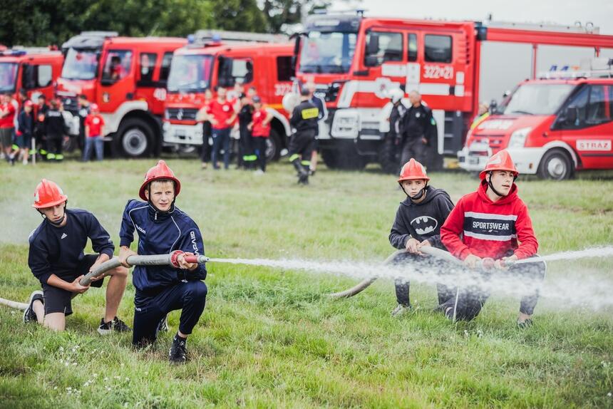 czterech nastolatków w strojach strażaków, po dwóch przy wężach strażackich z których leci woda, w przyklęku na trawie