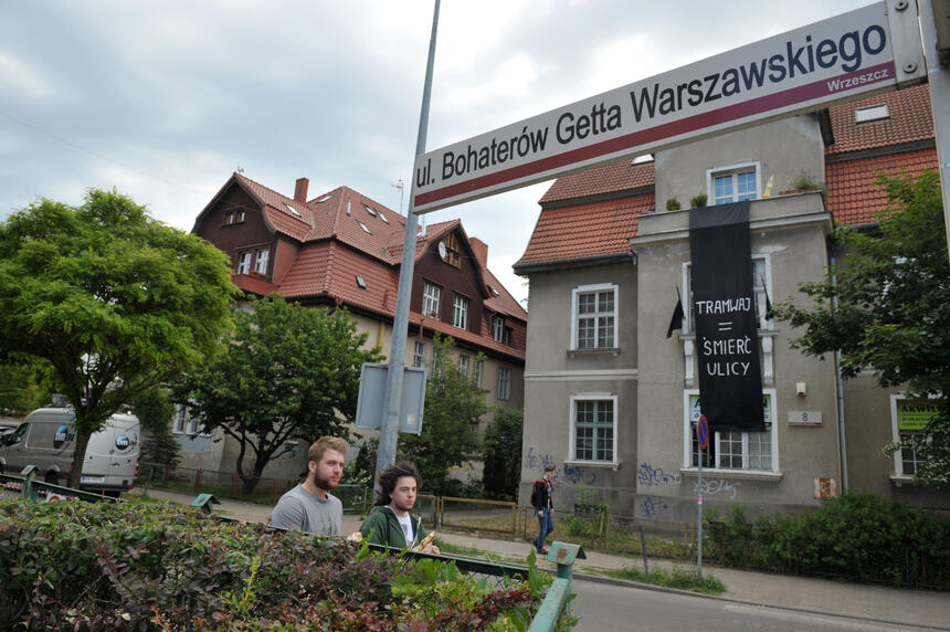 na zdjęciu na pierwszym planie tabliczka z nazwą ulicy Bohaterów Getta Warszawskiego, za nią widać do połowy dwóch idących młodych mężczyzn, w tle widać dwie charakterystyczne gdańskie kamienice