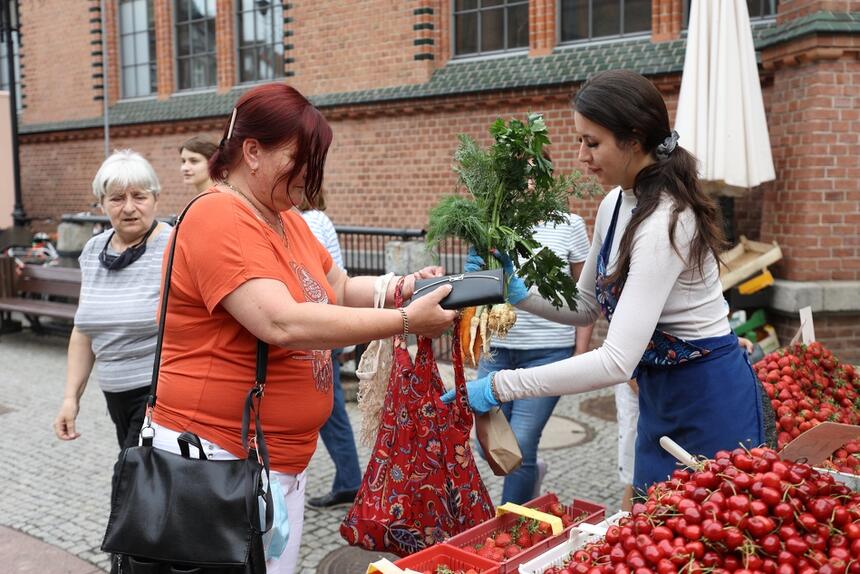 na zdjęciu młoda kobieta z lewej, sprzedawczyni, podaje zapakowane zakupy kobiecie z prawej strony, przed nimi na straganie leżą truskawki i czereśnie