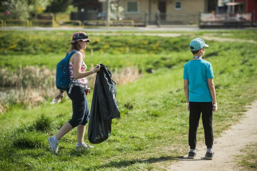 Dziewczynka i chłopiec na drodze, obok trawnik. Dziewczynka (po lewej) z czarnym workiem na śmieci