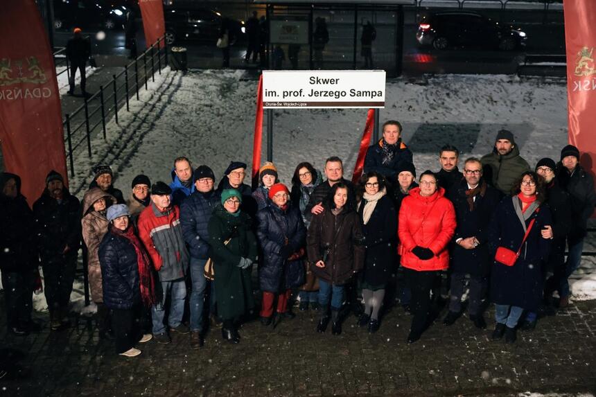 Grupa ponad dwudziestu osób sfotografowana wieczorową porą na tle białej tablicy z napisem Skwer im. prof. Jerzego Sampa 