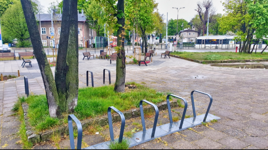 na zdjęciu fragment skweru, na pierwszym planie widać stojaki rowerowe, za nimi drzewo, w tle inne drzewa a także widać ławki, za nimi w tle widać dwie szare niewysokie kamienice i jadący kremowy tramwaj