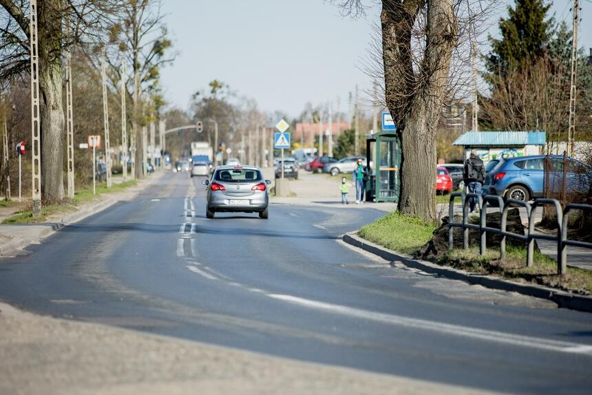 na zdjęciu fragment ulicy kielnieńskiej, widać jadący nią samochód osobowy, po prawej jest wiata przystankowa w której stoi kilka osób, wokół widać rosnące drzewa przy ulicy