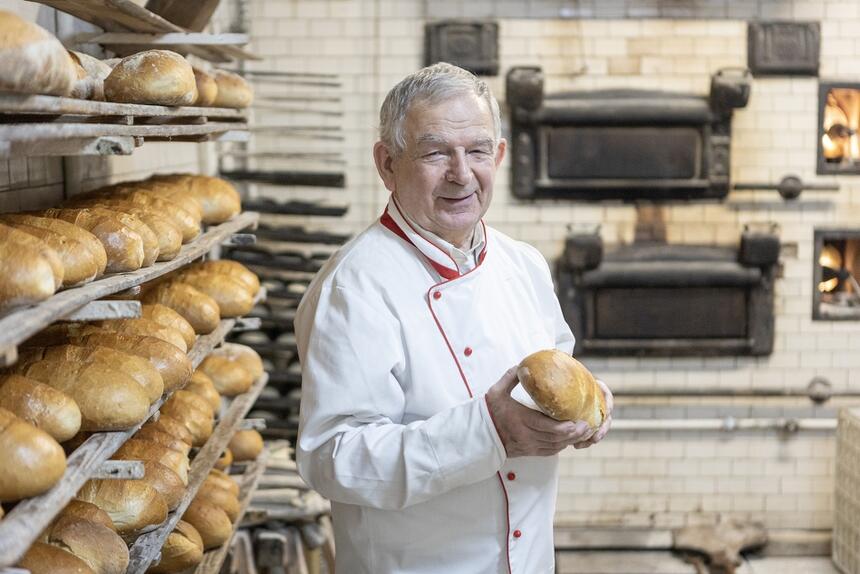 starszy mężczyzna w białym kitlu trzyma chleb, za nim stary kaflowy piec