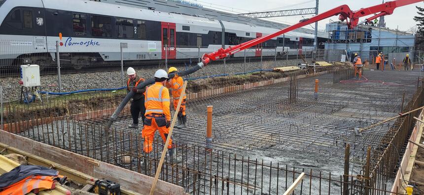 na zdjęciu plac budowy, widać betonowanie płyty fundamentowej, na której stoi kilku robotników w pomarańczowych kombinezonach, za nimi w tle przejeżdża szary pociąg