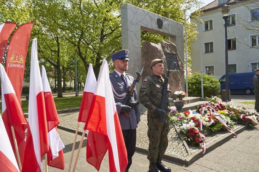 Po lewej flagi polskie i Gdańska, bok nich stoi dwóch żołnierzy, za nimi po prawej pomnik. W tle blok mieszkalny