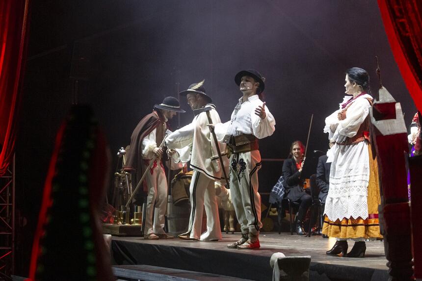 na scenie widać kilku mężczyzn w tradycyjnych strojach góralskich, występują na scenie