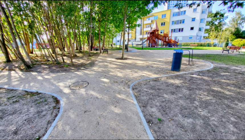 na zdjęciu fragment parku, widać wytyczone ścieżki spacerowe, rosnące zielone drzewa, kilka urządzeń zabawowych dla dzieci, po prawej fragment budynku
