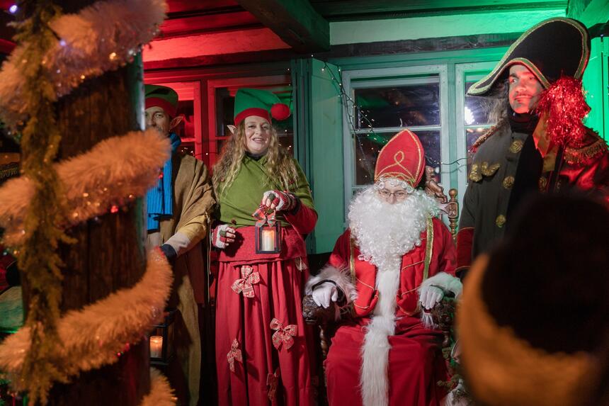na zdjęciu mikołaj w biało czerwonym stroju, siedzi na tronie, obok niego po lewej jest elfica w zielonym stroju, a po prawej stoi mężczyzna w wojskowym stroju sprzed dwustu lat