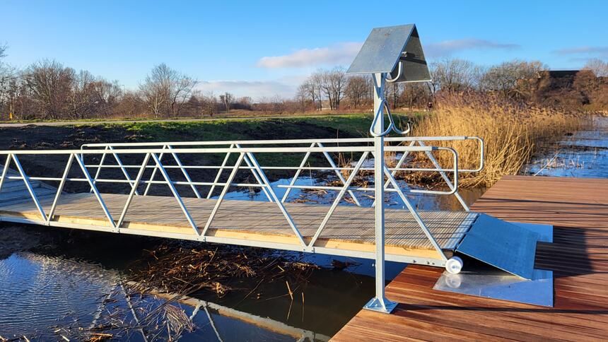 Zdjęcie przedstawia nowoczesny most pontonowy z metalową konstrukcją, który został zainstalowany nad niewielkim zbiornikiem wodnym. Konstrukcja mostu opiera się na geometrycznych, białych metalowych balustradach oraz brązowych, drewnianych deskach, które tworzą chodnik